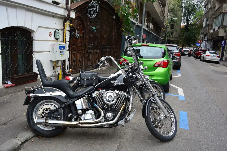 O motocicletă neagră cu elemente cromate este parcată lângă o mașină verde, pe o stradă mărginită de clădiri.
