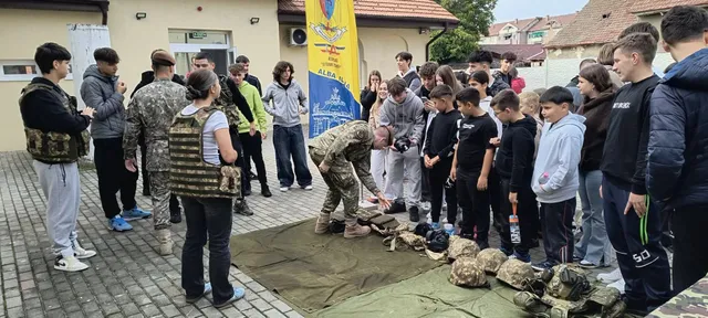 Doi oameni în uniforme de camuflaj stau lângă un grup de tineri; unul se apleacă spre echipamentele așezate pe jos.
