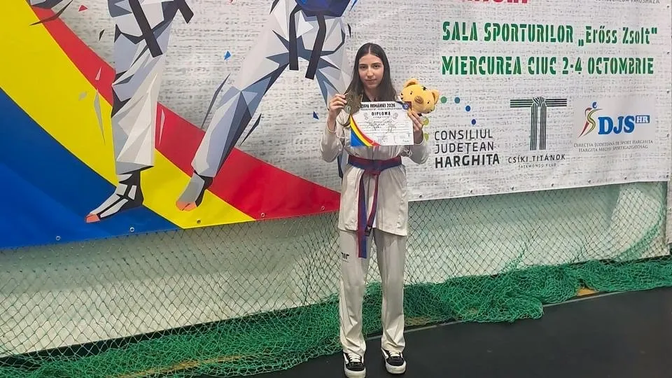 O sportivă în echipament alb ține o medalie, o diplomă și o jucărie de pluș, în fața unui panou sportiv.