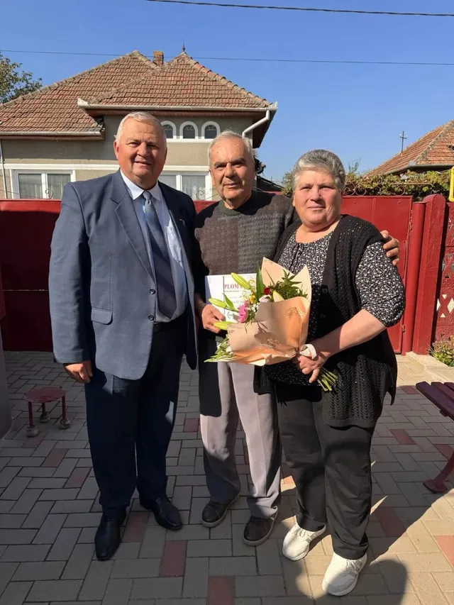 Trei adulți stau pe o alee pavată; bărbatul din mijloc și femeia din dreapta țin un buchet și o hârtie.