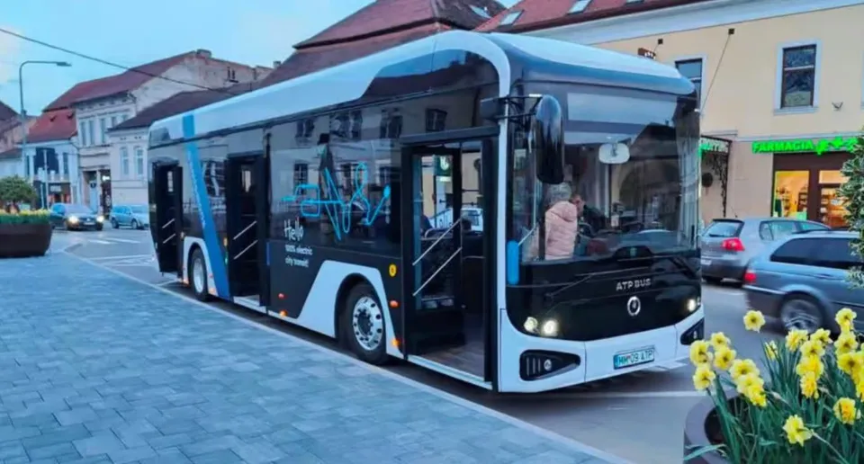 Un autobuz albastru și alb se află lângă trotuar, în fața unor clădiri; în prim-plan sunt flori galbene.