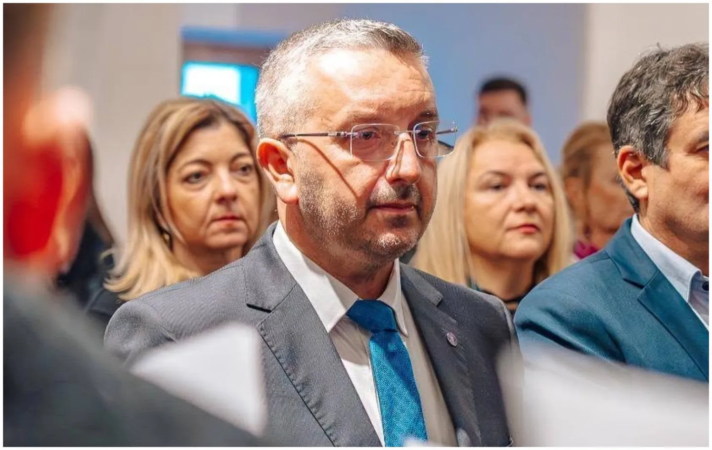 Un bărbat cu ochelari poartă sacou gri și cravată albastră, înconjurat de mai multe persoane, dintre care două femei sunt vizibile în spatele lui.