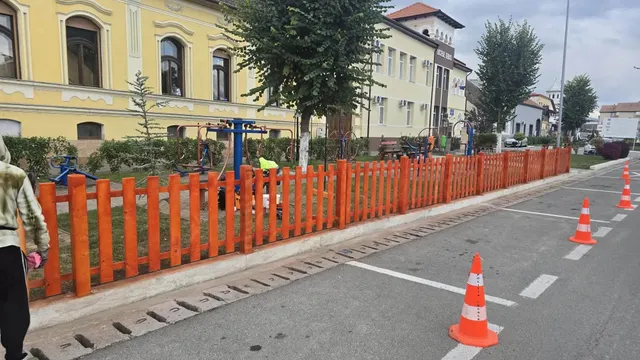 Un gard portocaliu mărginește o zonă cu iarbă și aparate, iar pe carosabil se văd conuri și marcaje albe.
