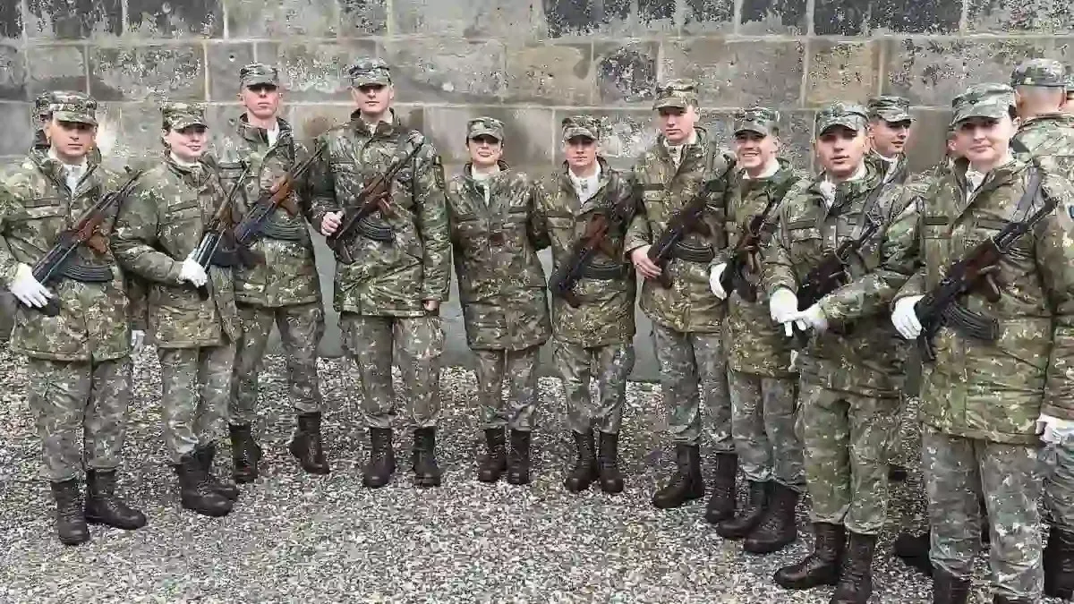 Un grup de persoane în uniforme de camuflaj stă aliniat în fața unui zid de piatră; unele poartă mănuși albe și țin arme.