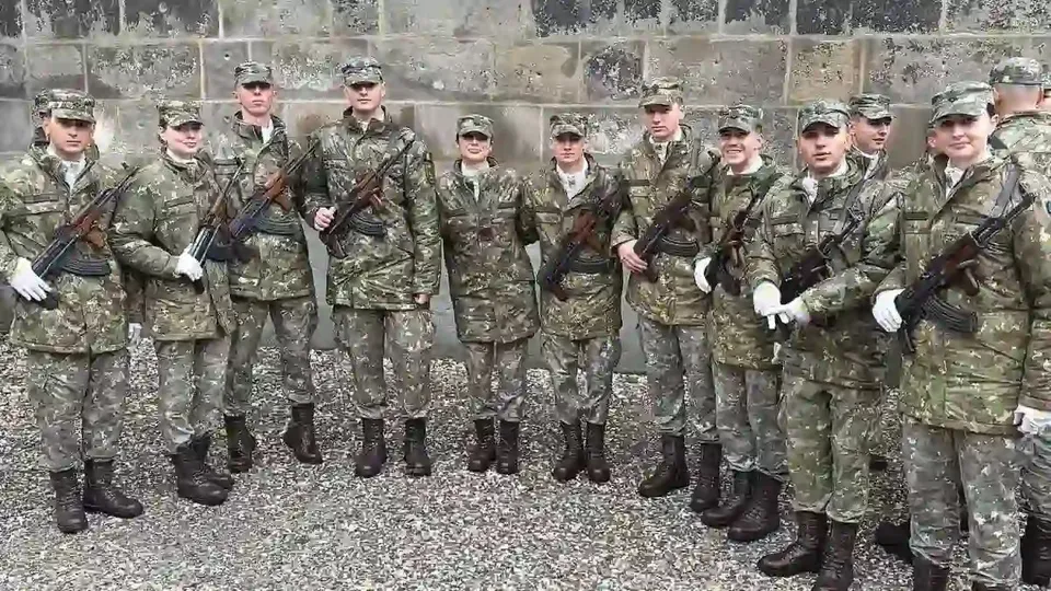 Un grup de persoane în uniforme de camuflaj stă aliniat în fața unui zid de piatră; unele poartă mănuși albe și țin arme.