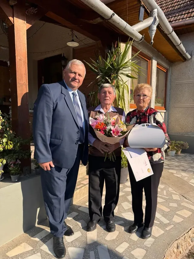Un bărbat în costum stă lângă două femei; una ține un buchet, iar cealaltă ține un obiect alb și o foaie.