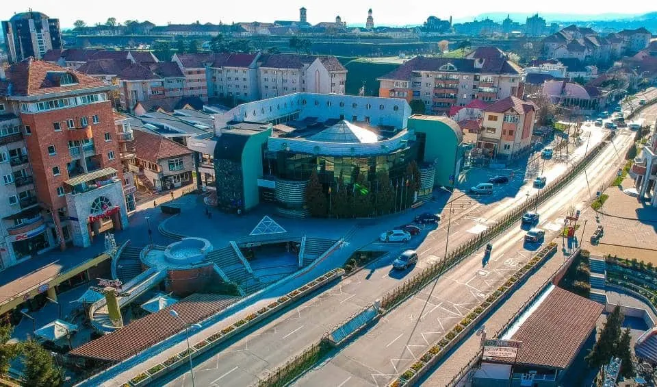 Vedere aeriană asupra unei zone urbane, cu clădiri, străzi, autoturisme, linii de transport și o construcție circulară în centru.