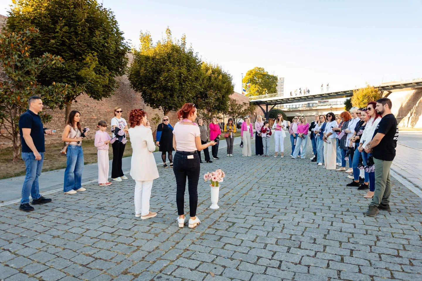 Mai multe persoane stau în formație curbată pe o alee pavată, unele ținând umbrele roz, iar în față se află flori.