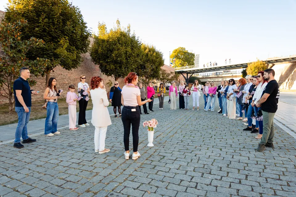Mai multe persoane stau în formație curbată pe o alee pavată, unele ținând umbrele roz, iar în față se află flori.