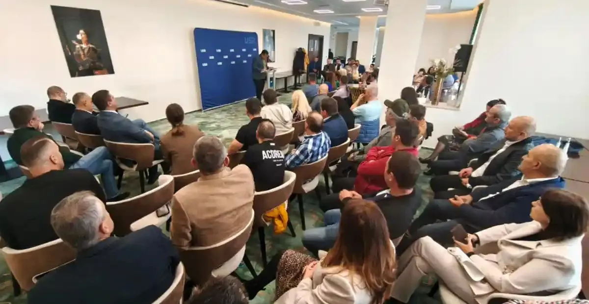 Mai multe persoane stau pe scaune într-o încăpere, orientate spre o masă și un panou albastru; câteva persoane stau în picioare lângă masă.