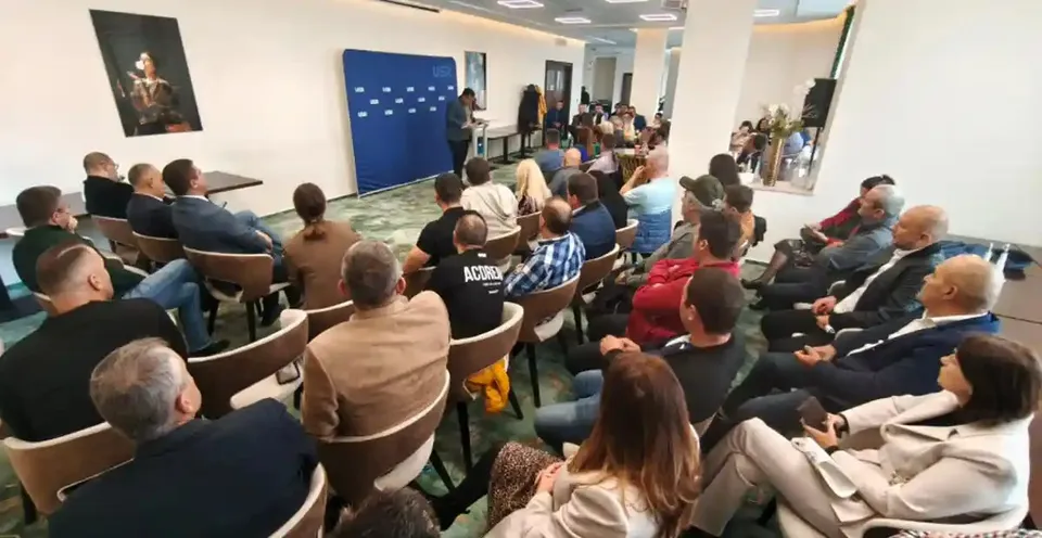 Mai multe persoane stau pe scaune într-o încăpere, orientate spre o masă și un panou albastru; câteva persoane stau în picioare lângă masă.