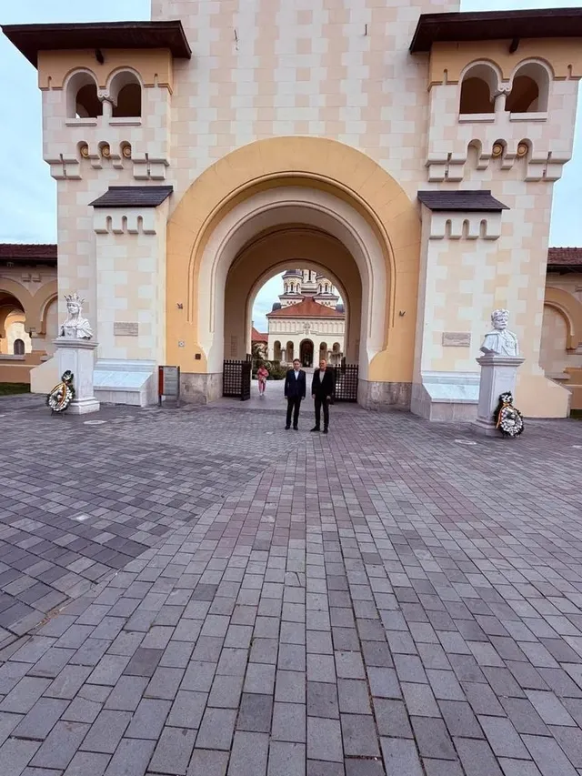 Două busturi albe sunt așezate de o parte și de alta a unei arcade, cu coroane de flori la bază; trei persoane stau sub arcadă.