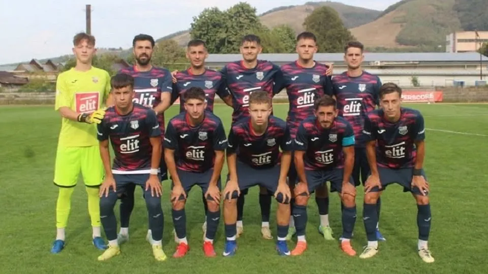 Unsprezece bărbați în echipament de fotbal pozează pe un teren cu iarbă; zece poartă tricouri închise la culoare, iar unul echipament galben.