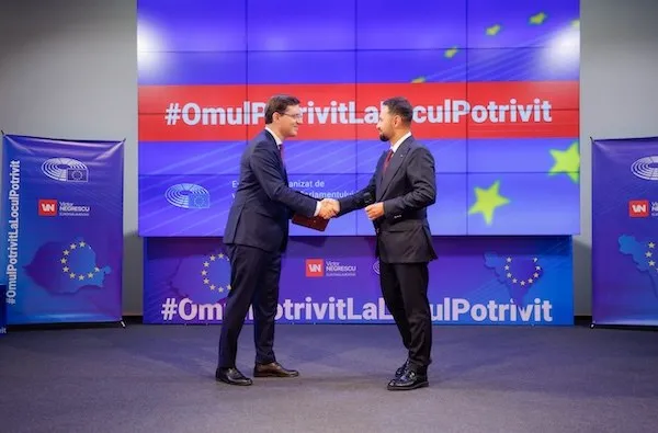 Doi bărbați în costume închise la culoare își strâng mâinile în fața unui panou violet cu text și stele; unul ține o hârtie.