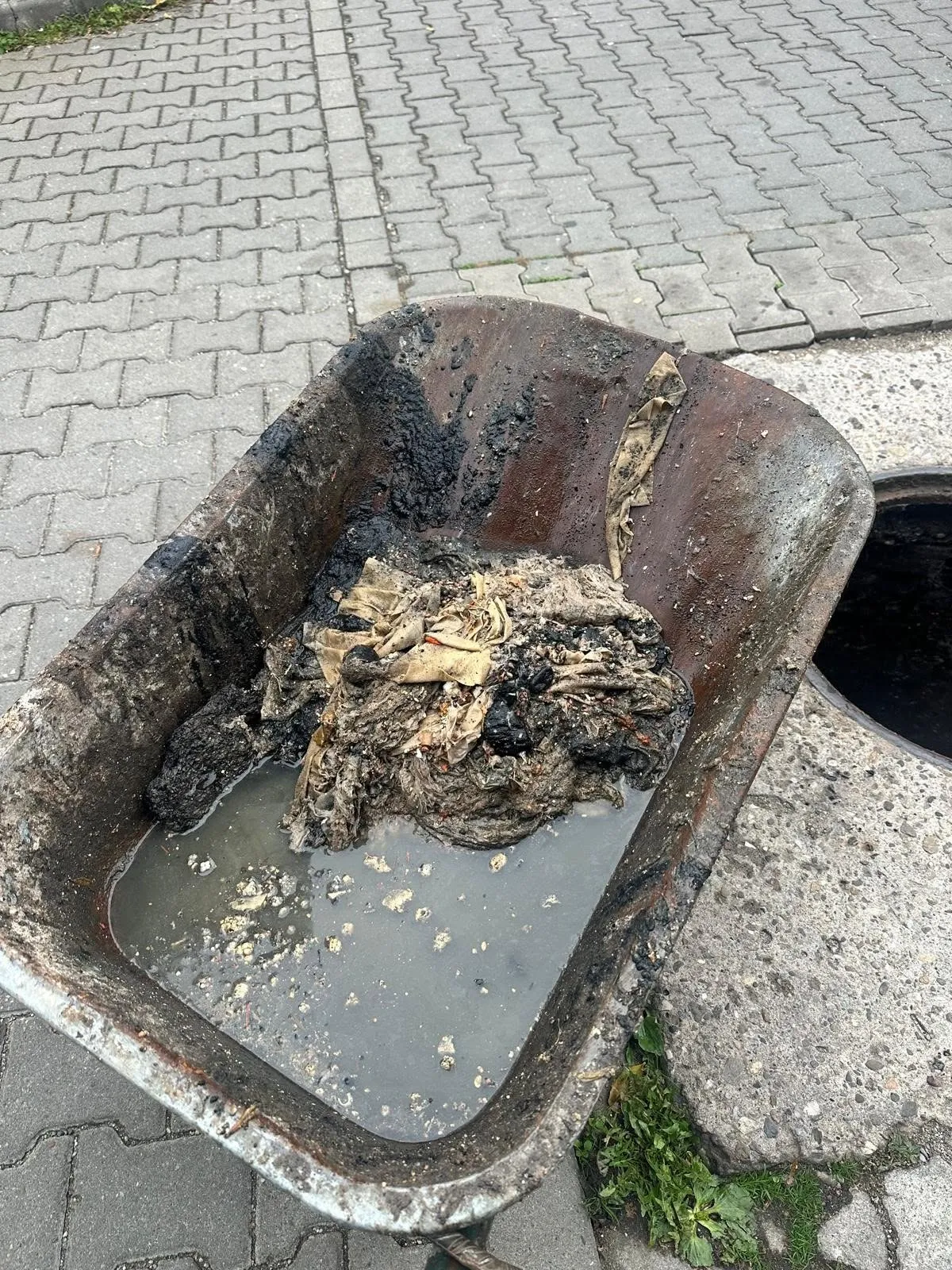 O cuvă metalică ruginită conține apă tulbure și o grămadă de materiale închise la culoare și deschise la culoare, lângă un pavaj.