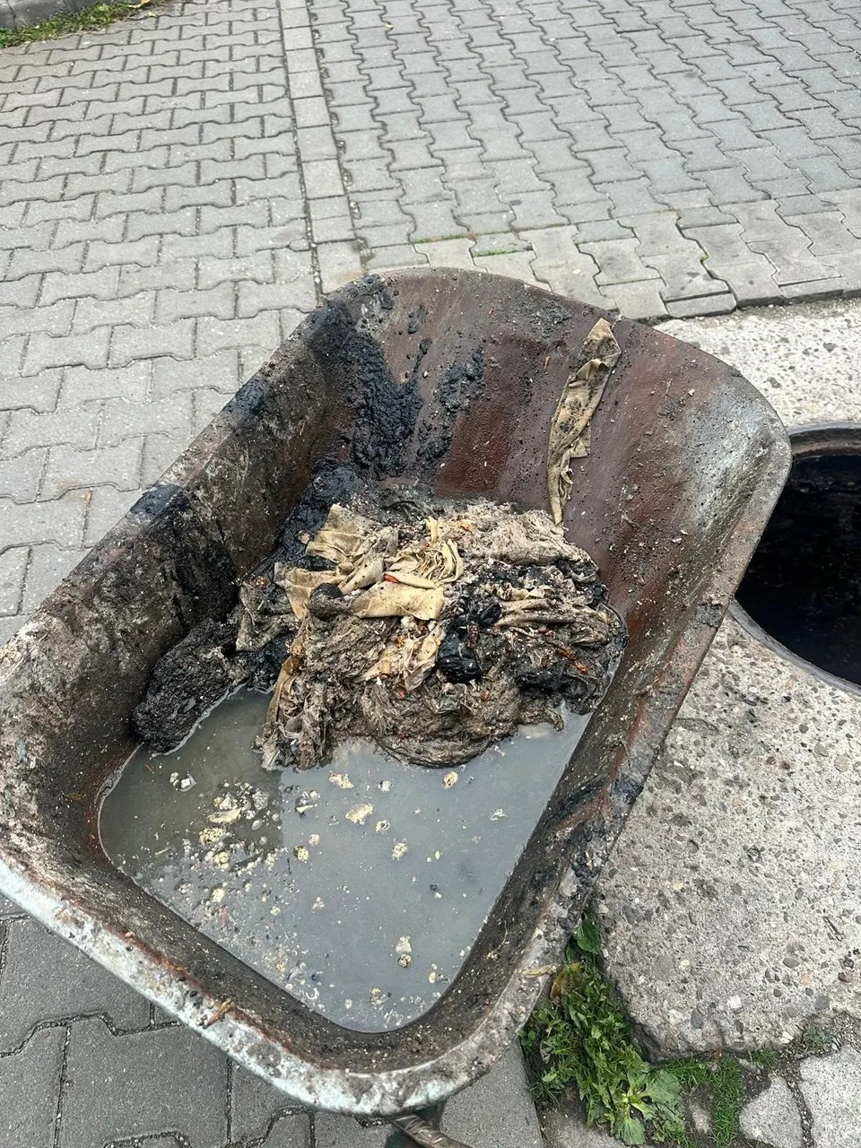 O cuvă metalică ruginită conține apă tulbure și o grămadă de materiale închise la culoare și deschise la culoare, lângă un pavaj.