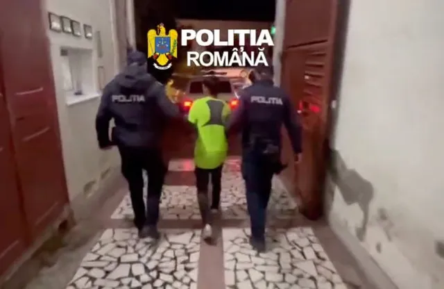 Doi oameni în uniforme închise la culoare merg de o parte și de alta a unei persoane în vestă verde, pe o alee pavată.
