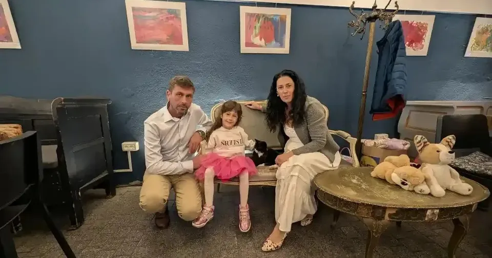 Un bărbat, o fetiță și o femeie stau pe scaune, lângă o masă cu jucării de pluș și picturi înrămate pe perete.