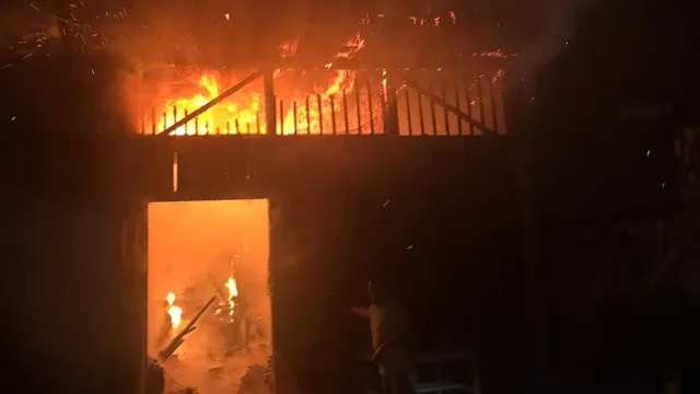 Flăcări portocalii se văd în spatele unui gard cu bare verticale, deasupra unei deschideri luminate; restul cadrului este întunecat.