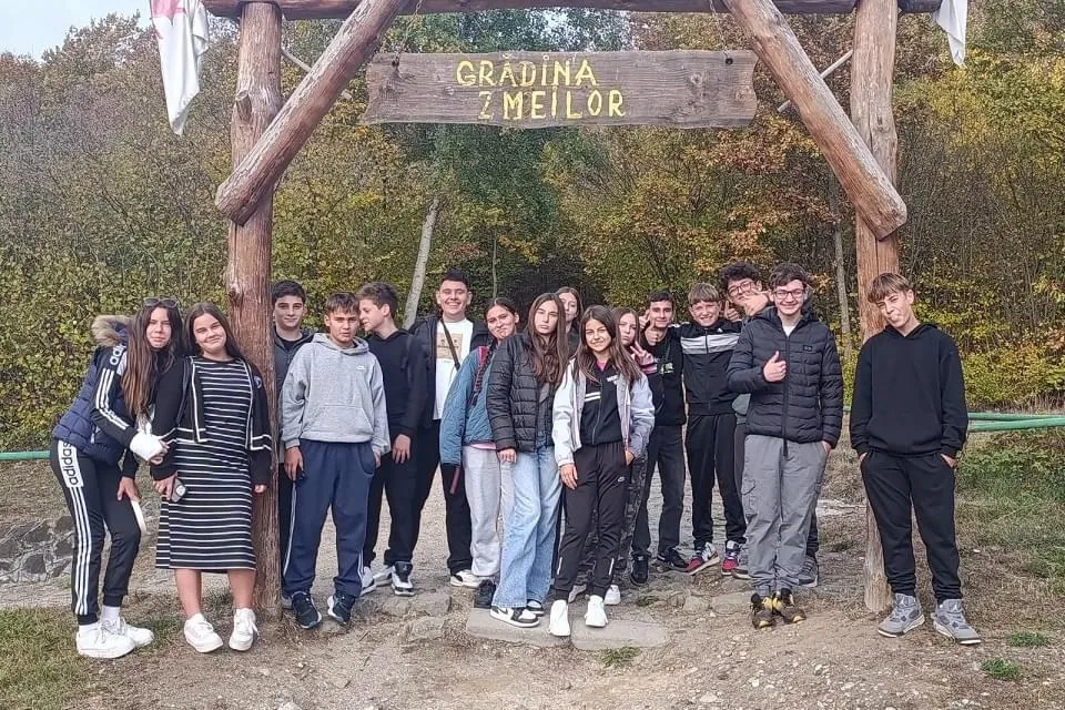 Un grup de tineri stă sub o poartă din lemn cu inscripția „GRĂDINA ZMEILOR”, pe o alee mărginită de copaci.
