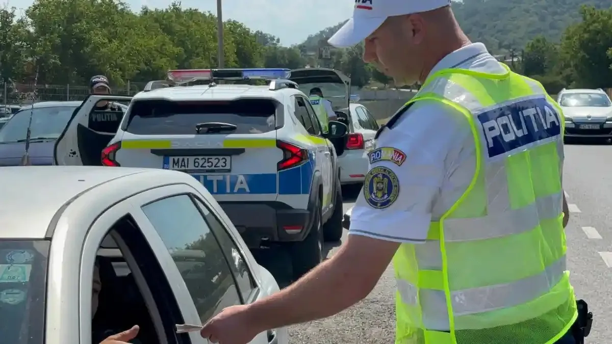 Un bărbat în vestă reflectorizantă și șapcă albă stă lângă un autoturism, cu brațul întins spre geamul deschis; în spate sunt autospeciale.