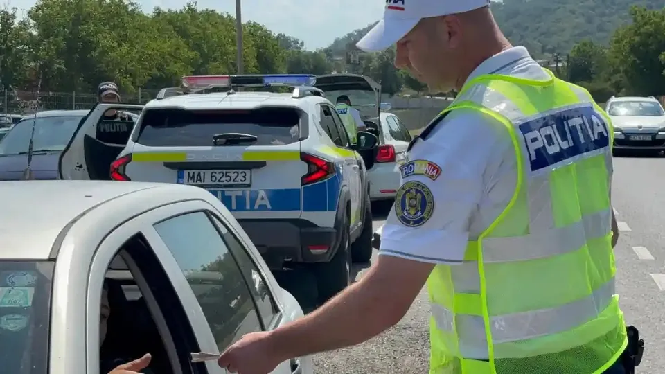 Un bărbat în vestă reflectorizantă și șapcă albă stă lângă un autoturism, cu brațul întins spre geamul deschis; în spate sunt autospeciale.