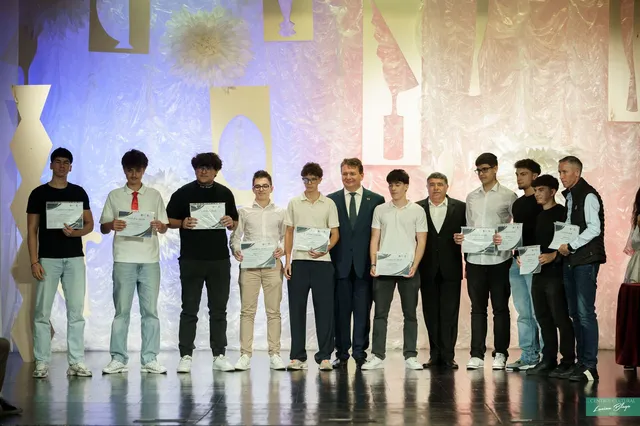 Un grup de persoane stă pe o scenă, majoritatea ținând diplome, în fața unui decor colorat.