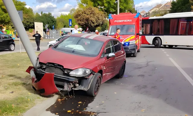 Un autoturism roșu cu partea din față avariată este lângă un stâlp, iar în spate se văd o autospecială roșie, un autobuz și câteva persoane.