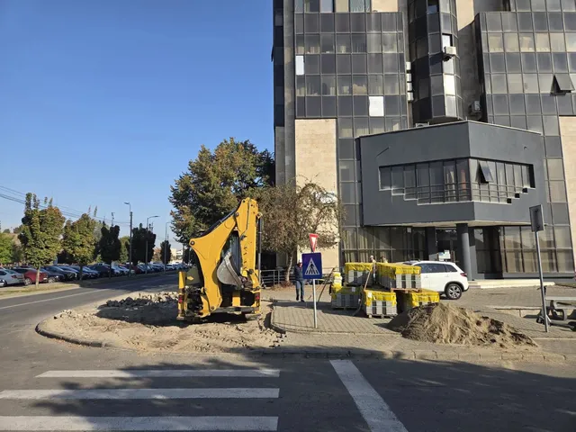 Un excavator galben sapă lângă o trecere de pietoni, iar un camion alb-galben și bariere galbene se află în apropiere.