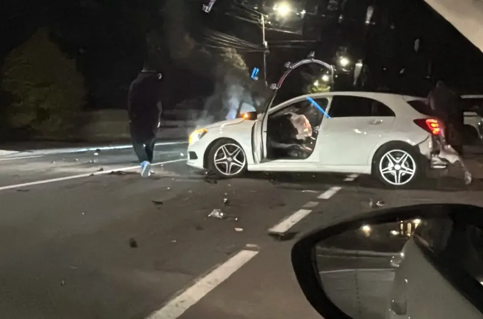 Un autoturism alb este oprit pe carosabil, cu portiera deschisă și partea din spate avariată; fragmente sunt împrăștiate, iar o persoană stă lângă el.