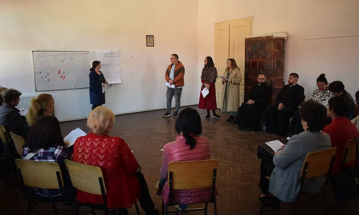 Mai multe persoane stau pe scaune dispuse în semicerc, iar patru persoane se află în picioare lângă o tablă și un dulap.