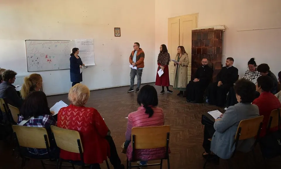 Mai multe persoane stau pe scaune dispuse în semicerc, iar patru persoane se află în picioare lângă o tablă și un dulap.
