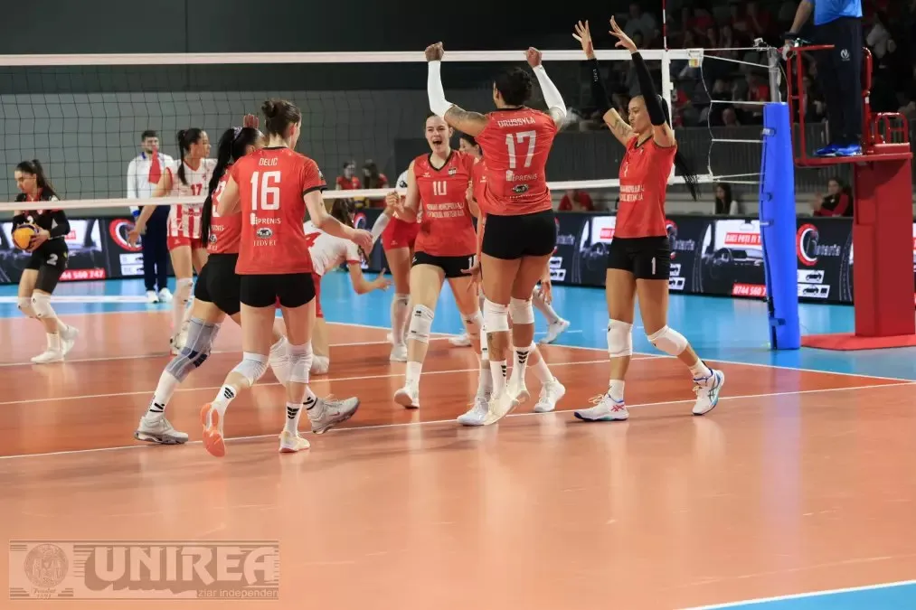 Pe un teren de volei, mai multe sportive în echipament roșu stau lângă fileu, unele cu brațele ridicate; o minge se vede în fundal.