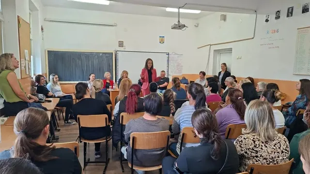 Într-o sală cu tablă și bănci, mai multe persoane stau pe scaune, iar o femeie aflată în picioare este aproape de mijlocul încăperii.
