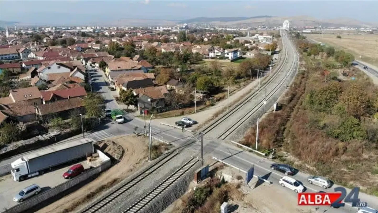 O vedere aeriană arată șine de cale ferată, un drum care le traversează, câteva vehicule, case și terenuri cu vegetație.