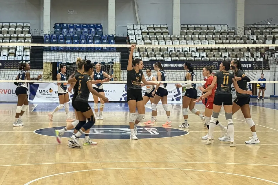 Jucătoare în echipamente negre, albe și roșii se află pe un teren de volei, de o parte și de alta a fileului.