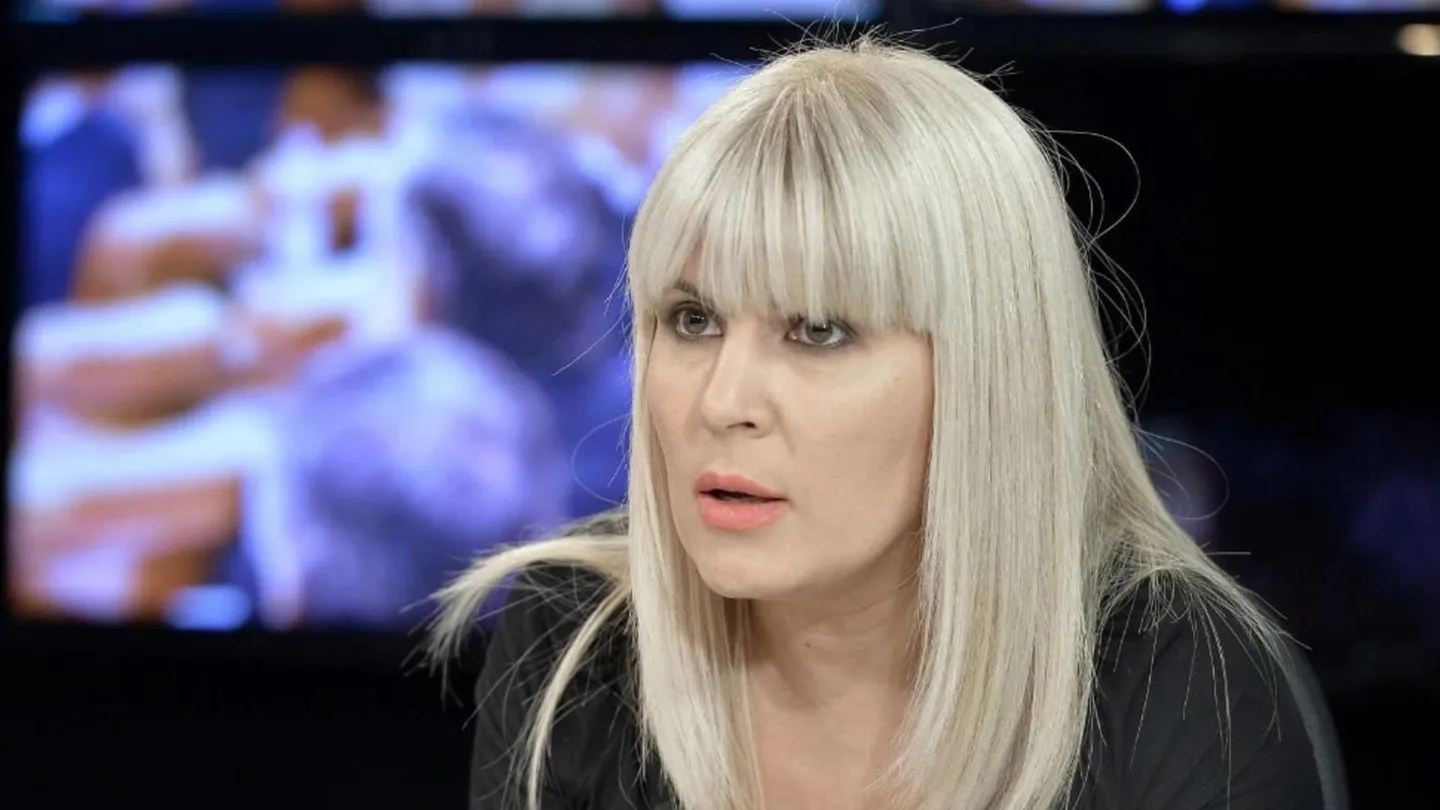 O femeie cu păr blond și breton poartă o haină neagră; în spatele ei se vede un ecran cu imagini neclare.