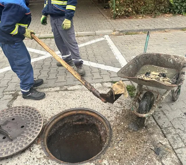 Două persoane în haine de lucru stau lângă un cămin deschis; una ține o lopată, iar alături se află o roabă cu materiale.