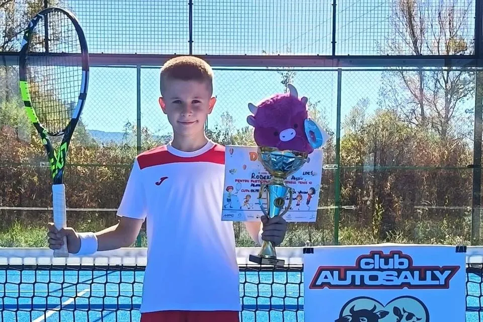 Un băiat în tricou alb-roșu ține o rachetă de tenis, o diplomă și un trofeu, lângă un panou și un fileu.
