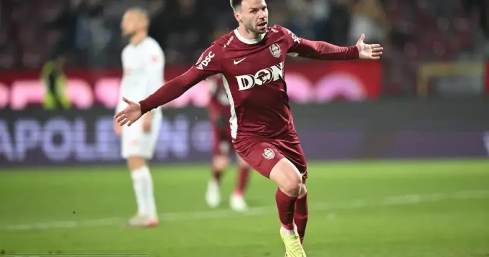 Un fotbalist în echipament vișiniu aleargă cu brațele întinse pe gazon; în spate se văd jucători în echipamente albe.