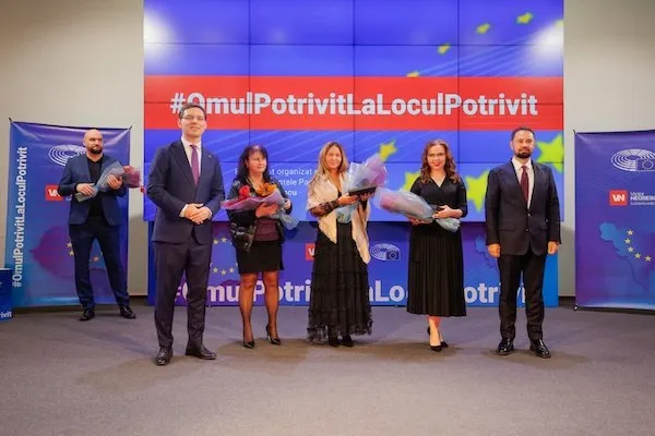 Șase persoane stau în picioare pe o scenă, în fața unui ecran cu text; trei țin buchete de flori.