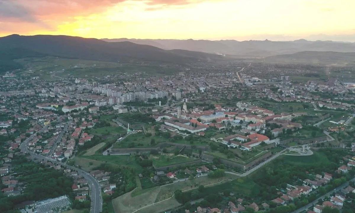Vedere aeriană asupra unui oraș, cu fortificații geometrice, clădiri cu acoperișuri roșii, zone verzi, drumuri și munți la orizont.