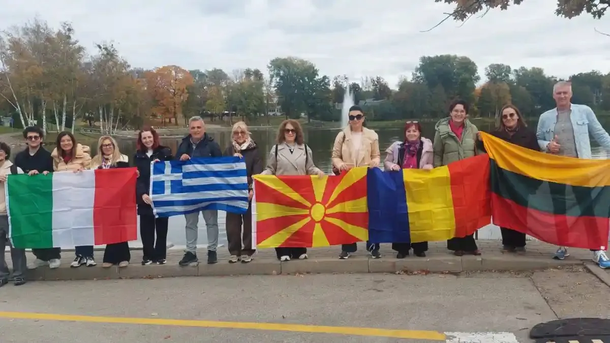 Un grup de persoane stă lângă un drum și ține steaguri multicolore; în spate se văd copaci și un gard.