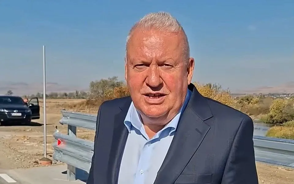 Un bărbat cu păr cărunt, în sacou închis și cămașă deschisă, stă în fața unei bariere metalice; în stânga se văd mașini parcate.