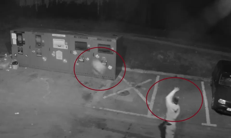 Imagine alb-negru cu mai multe containere metalice, o persoană cu un braț ridicat, un autoturism parțial vizibil și marcaje pe asfalt.