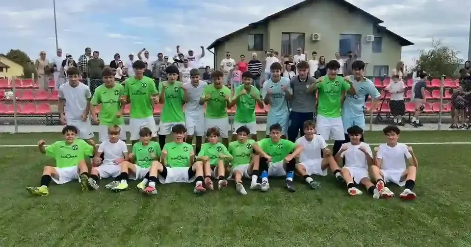 Un grup de tineri în tricouri verzi sau albe pozează pe iarbă, unii așezați, alții în picioare, cu tribune și o clădire în spate.