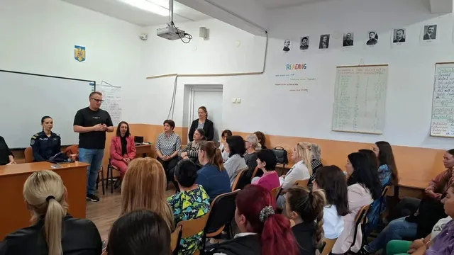 Un bărbat în picioare ține un obiect în mâini, în fața unor persoane așezate pe scaune într-o sală.