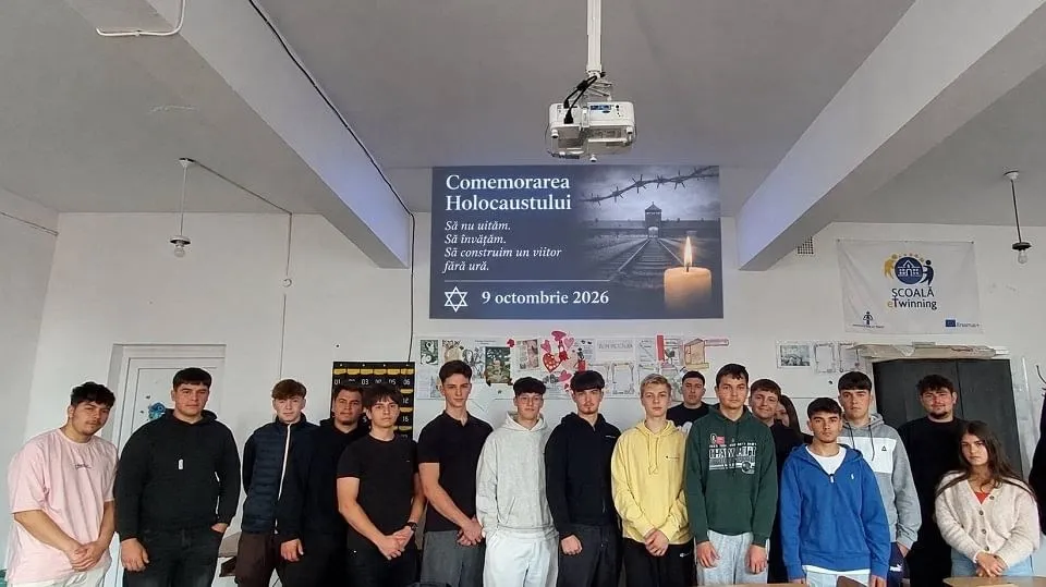 Un grup de persoane stă în fața unei table și a unui afiș, într-o încăpere cu tavan alb și videoproiector.