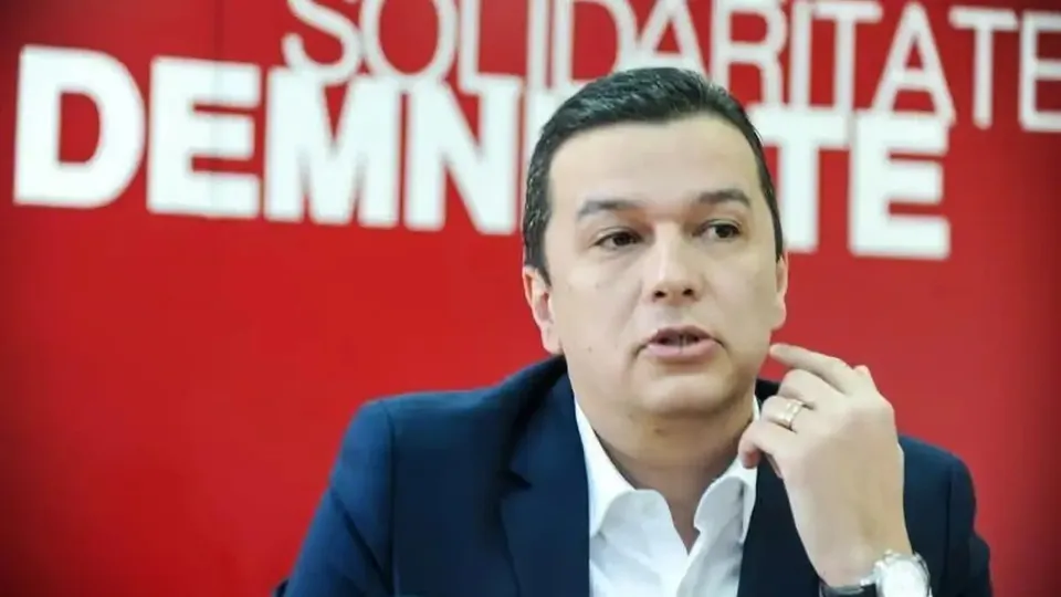 Un bărbat cu păr scurt, îmbrăcat într-un sacou închis la culoare și o cămașă albă, ține o mână ridicată lângă față, în fața unui fundal roșu cu litere albe.