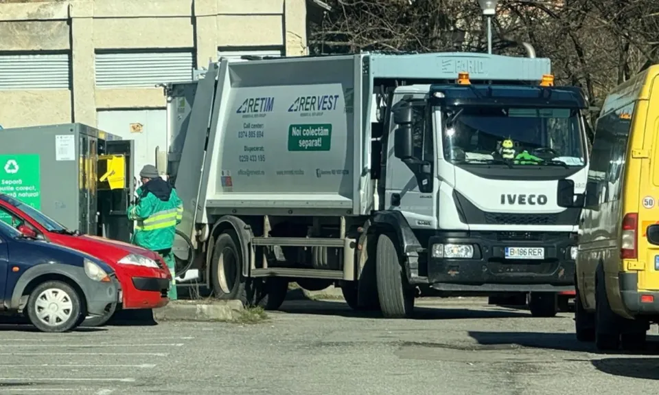 O autospecială albă de colectare a deșeurilor este parcată lângă o clădire; în apropiere se află un lucrător cu vestă verde.